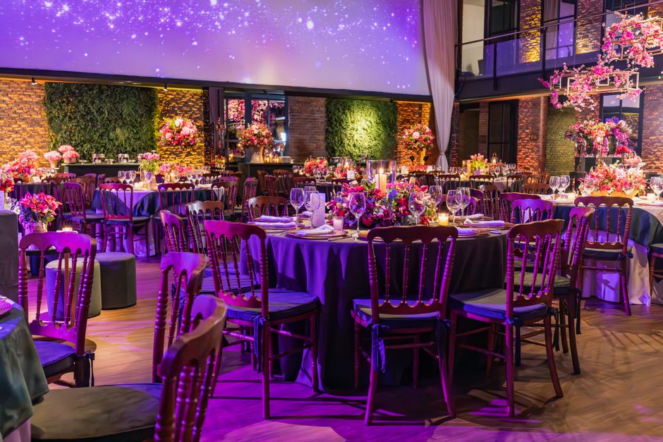 An elegantly arranged event space featuring multiple round tables covered with deep purple tablecloths, each adorned with vibrant floral centrepieces consisting of pink, purple, and white blossoms. Th