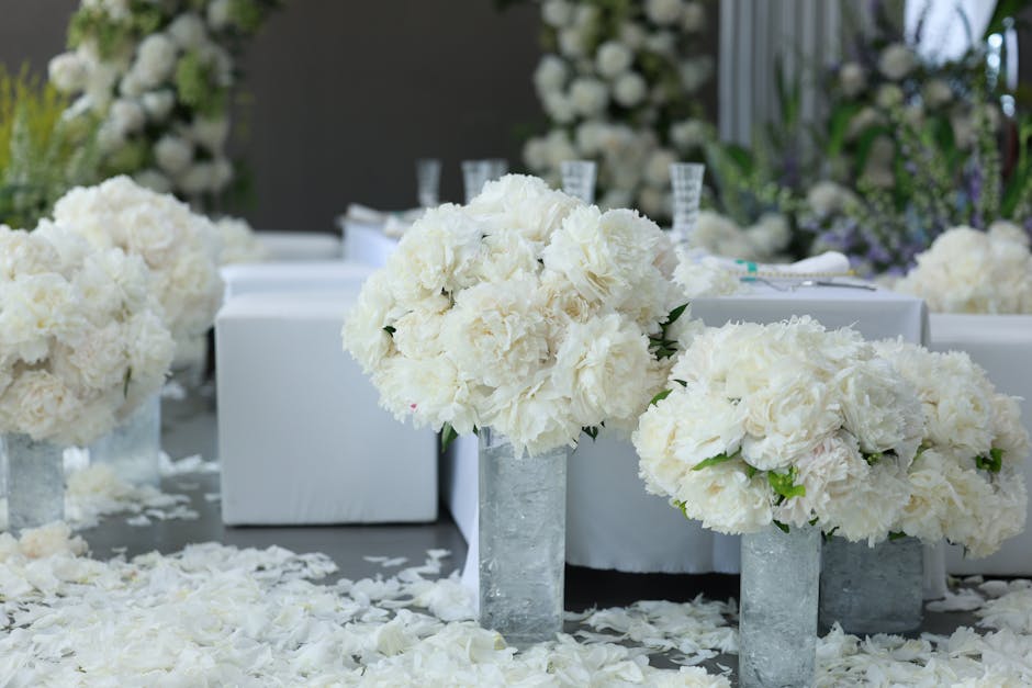 A floral arrangement featuring large bouquets of white hydrangeas and roses, arranged in tall, cylindrical glass vases placed on a white table. The flowers are fresh with soft, textured petals and gre