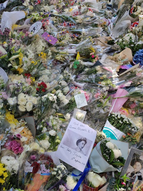 A large, somber display of numerous floral arrangements and bouquets laid out on the ground, predominantly featuring white, purple, and pink flowers such as roses, carnations, and chrysanthemums. Many