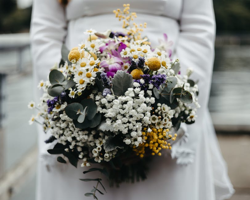 A person dressed in white holding a large, lush bouquet of fresh flowers outdoors. The bouquet features a mix of white daisies, small white baby's breath, yellow billy balls, purple flowers, and silve