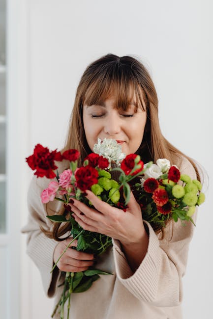 A young woman with light brown hair and bangs, dressed in a light beige sweater, gently holds a large bouquet of fresh flowers close to her face with her eyes closed and a content expression. The bouq