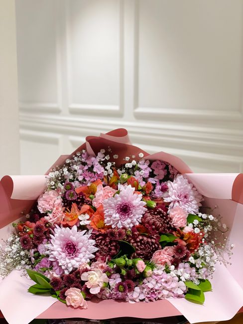 A vibrant floral bouquet featuring a mix of fresh pink and purple chrysanthemums, dahlias, and smaller filler flowers such as baby's breath, arranged in a lush and full style. The bouquet includes gre
