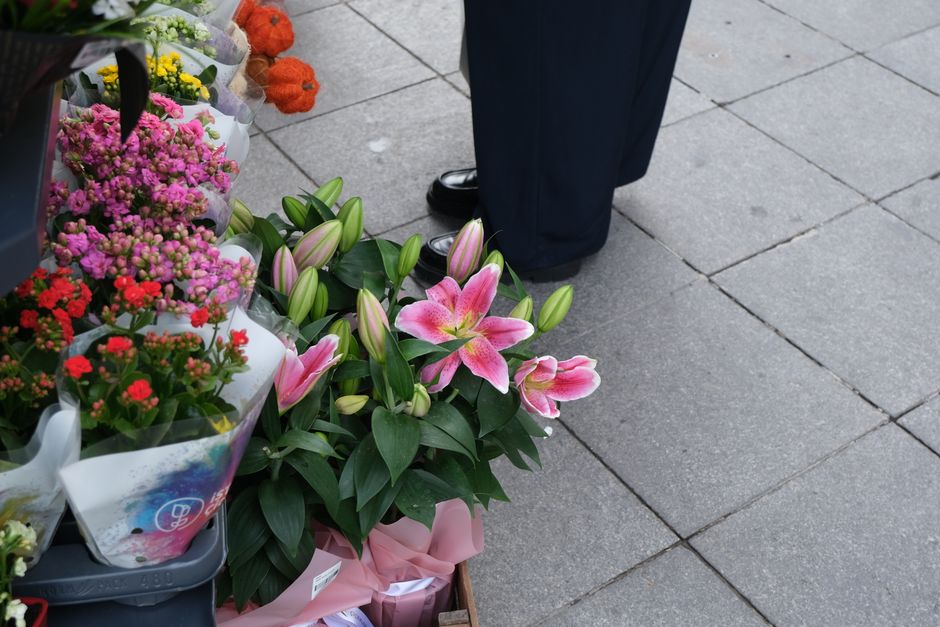 A floral arrangement featuring pink lilies with open blooms and partially closed buds, arranged alongside glossy green leaves. The lilies are placed on a sidewalk near other bouquets wrapped in pink t