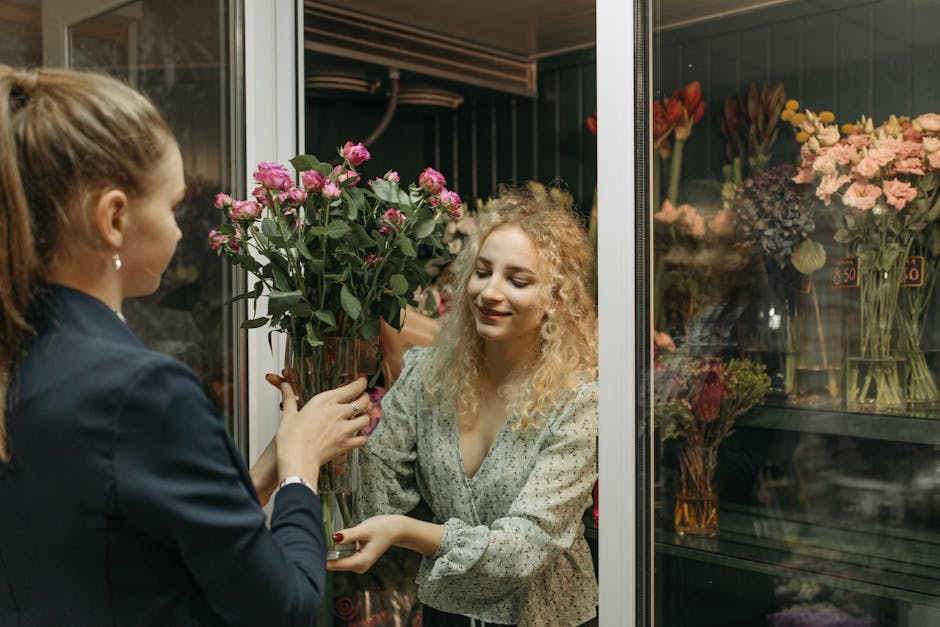 A female florist with curly blonde hair, dressed in a light patterned blouse, is handing a tall, clear glass vase filled with fresh pink roses to a customer with long brown hair tied back, wearing a d