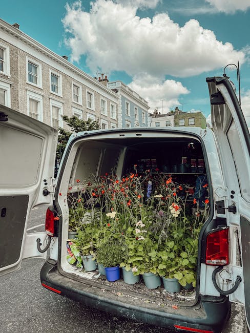 The open rear of a white delivery van parked on a street reveals an array of fresh flowering plants, including red and white flowers with green foliage, arranged in small blue and gray pots. The van's